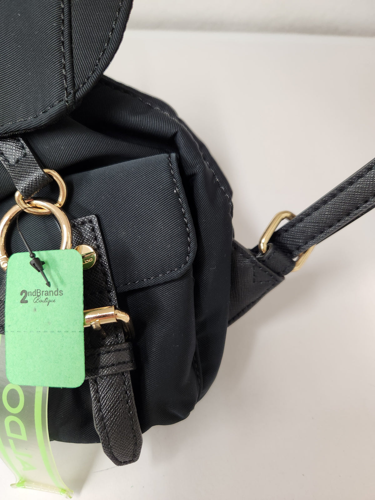A close-up of a black nylon shoulder bag with gold-tone hardware, featuring a green paper tag from 2Binds Studio attached.