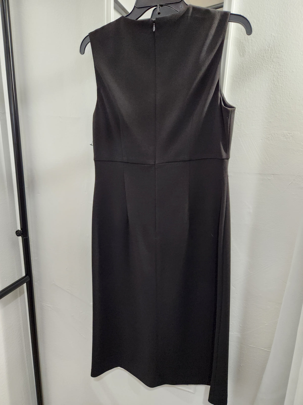 A sleeveless black midi dress hanging on a black hanger against a white curtain backdrop.