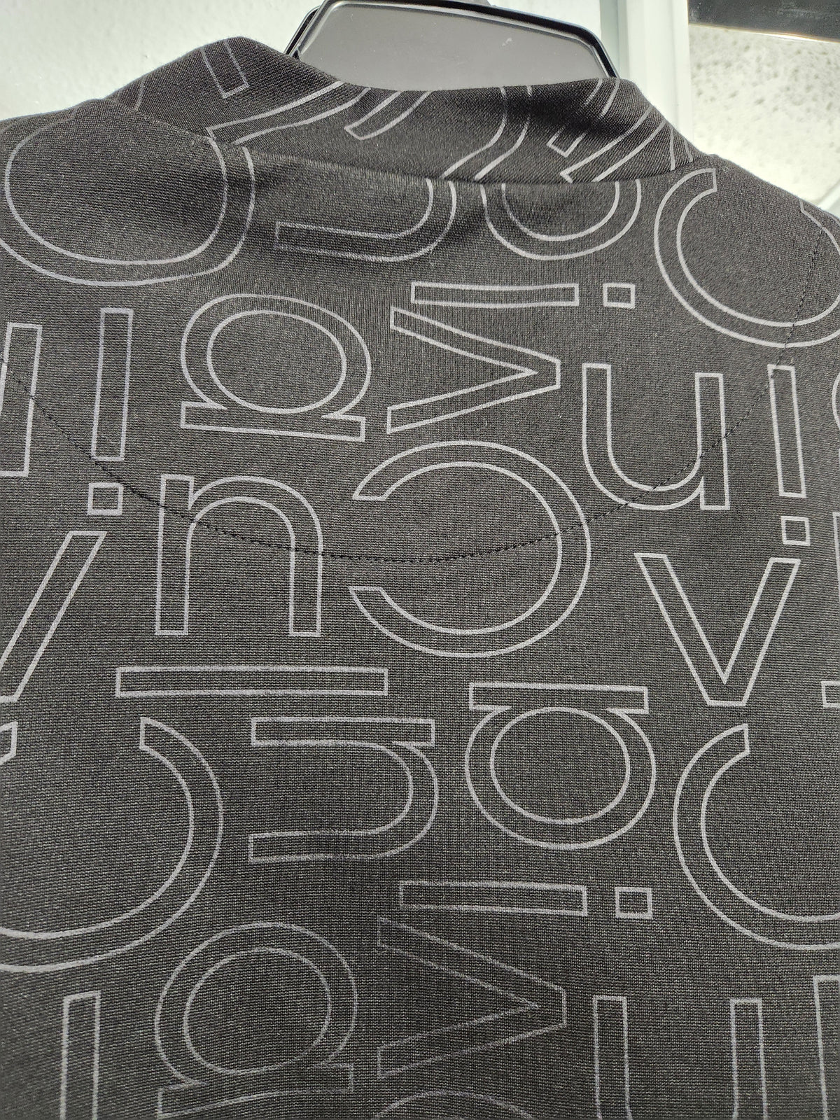 Close-up of a dark gray sweatshirt featuring an all-over white outline pattern of stylized letters, shown on a hanger.