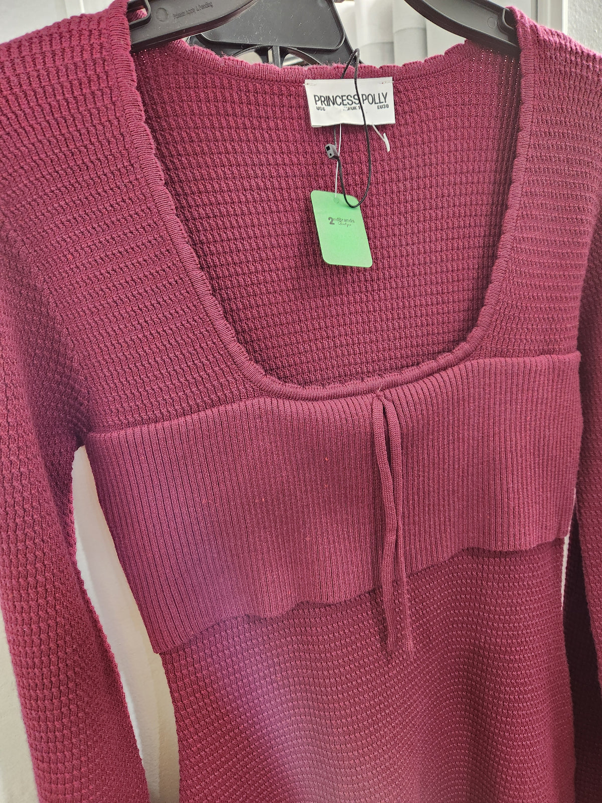 A close-up of a pink ribbed knit sweater hanging on a black hanger, featuring a scoop neckline, a textured horizontal band with a small front tie detail, and a visible Princess Polly brand tag.