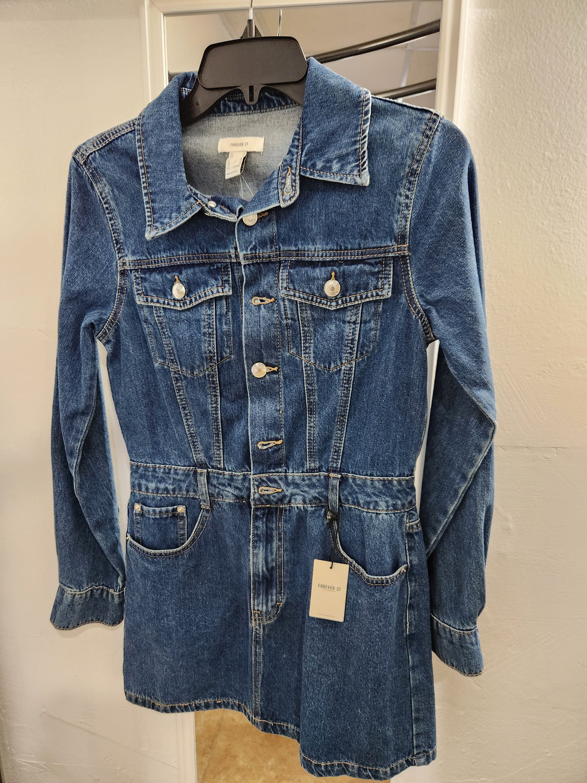 A blue denim long-sleeve mini dress with a button-front closure, chest flap pockets, and a self-tie belt, hanging on a black hanger against a white door.