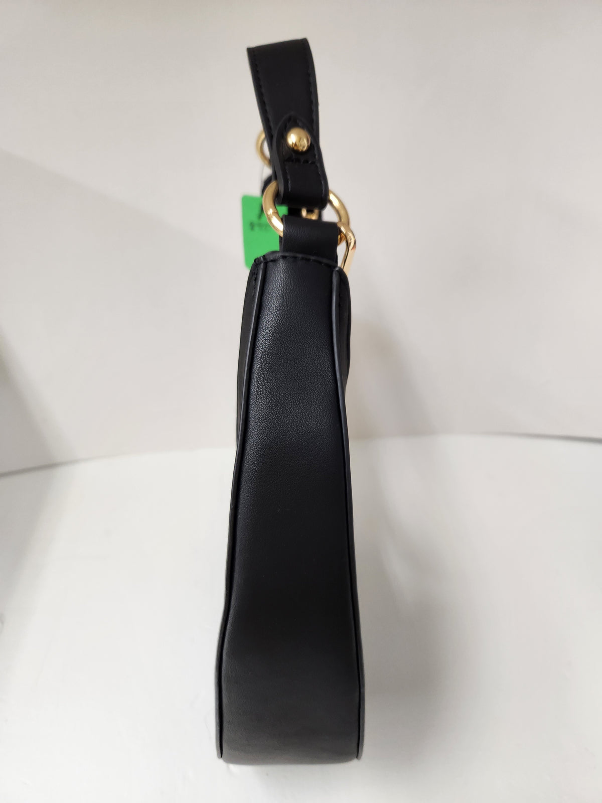A black faux-leather shoulder bag shown from a side profile view, featuring gold-tone hardware and a small green tag attached near the strap.