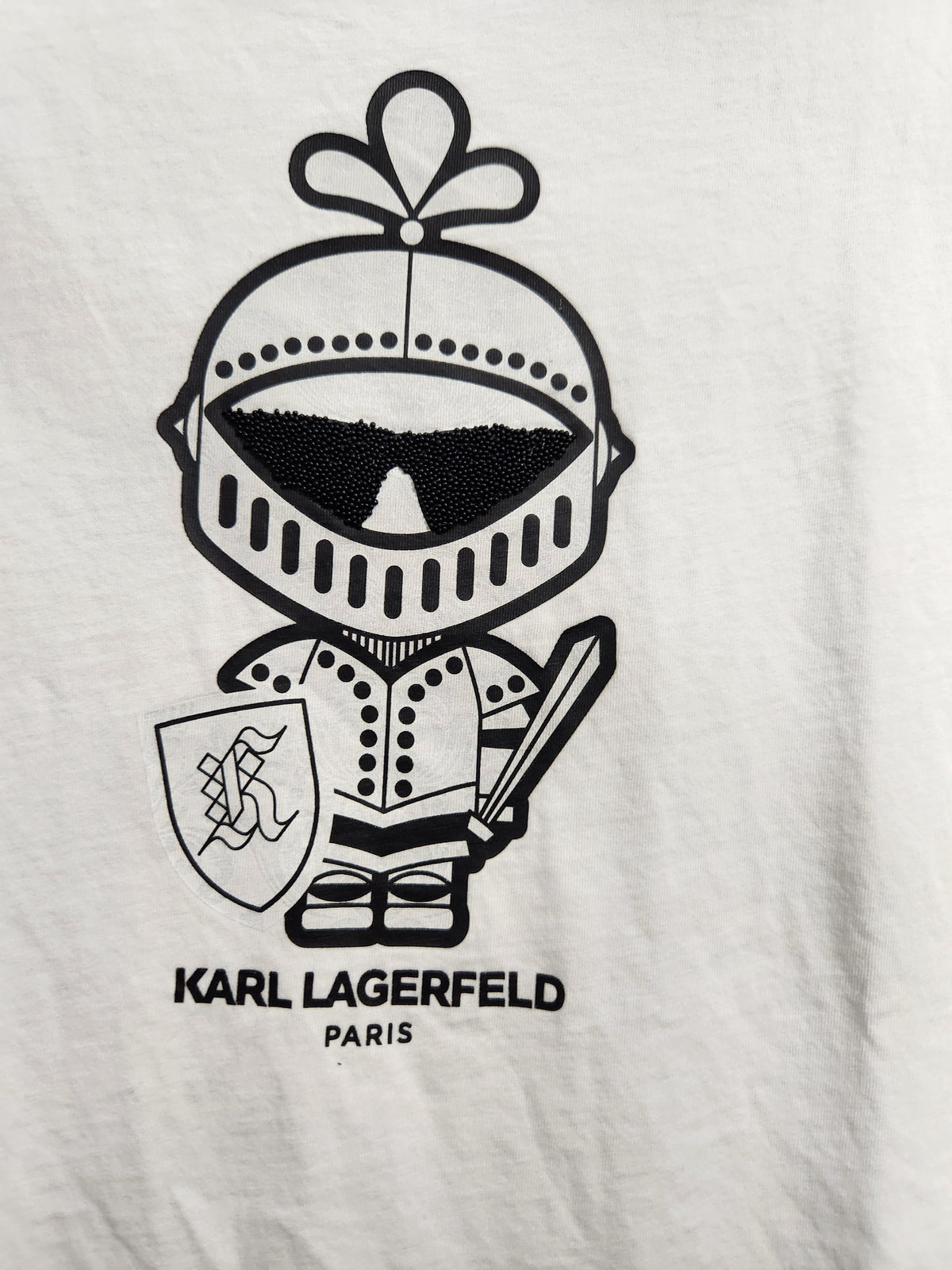 A close-up of a white fabric surface featuring a black graphic of a cartoon knight in armor with a helmet, shield, and sword, with the text 'KARL LAGERFELD PARIS' printed below.