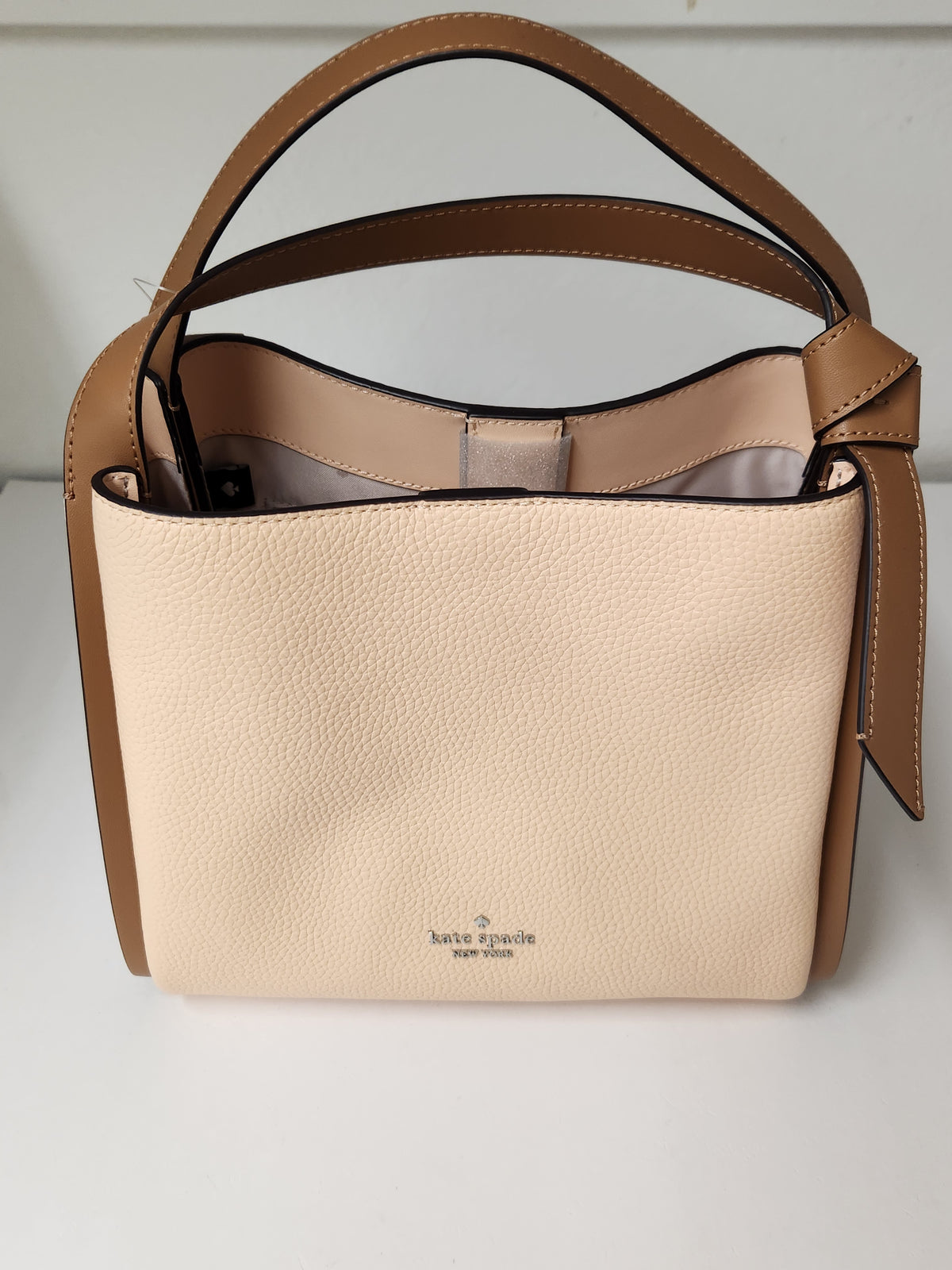 A structured pebbled leather handbag in beige and tan, featuring a top handle, a detachable shoulder strap, and a black hangtag on the side, photographed against a neutral light gray background.
