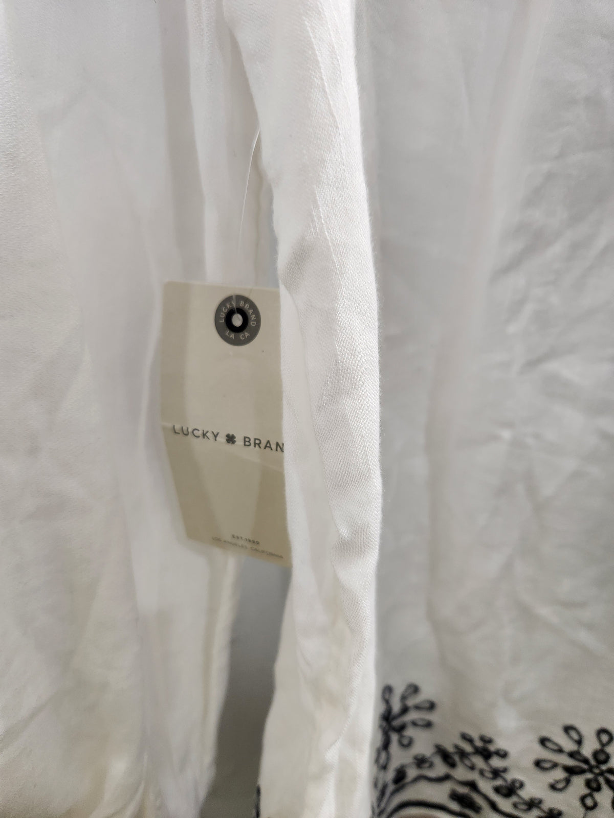 Close-up of a white textured fabric garment with a hanging paper tag reading 'Lucky Brand' and a small black embroidered floral motif near the bottom right.