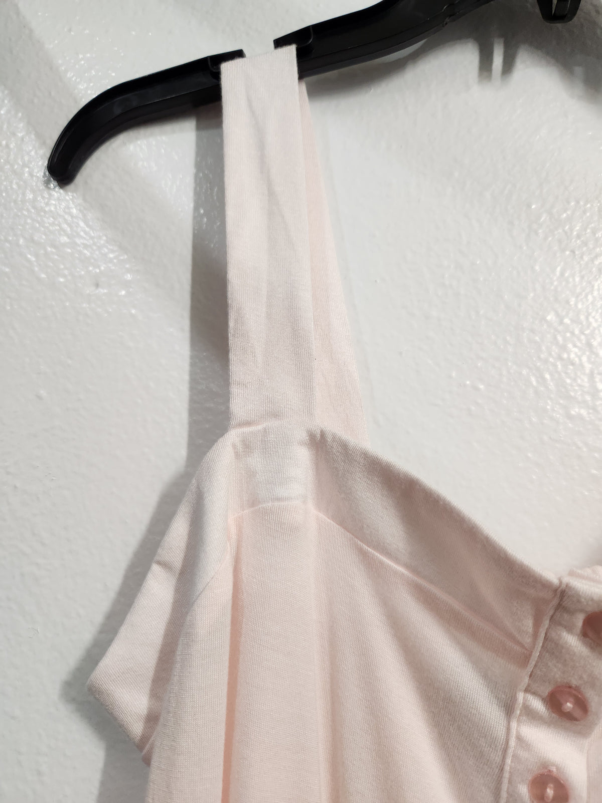 A close-up of a light blush pink sleeveless top hanging on a black hanger against a textured white wall. The top features a wide shoulder strap and a side button detail with pink buttons.