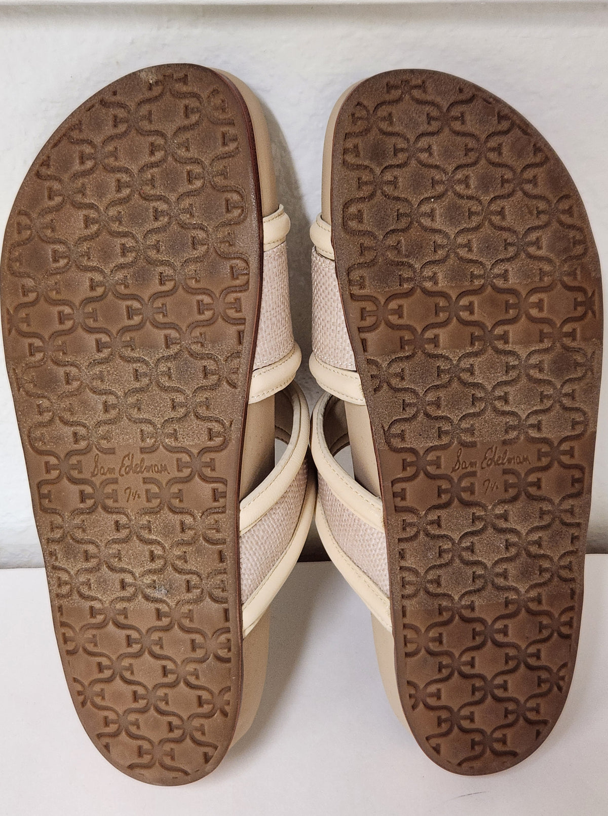 A pair of beige strappy sandals shown from the bottom, displaying the brown textured outsole with embossed interlocking 'C' logos and 'Joe Collins' branding.