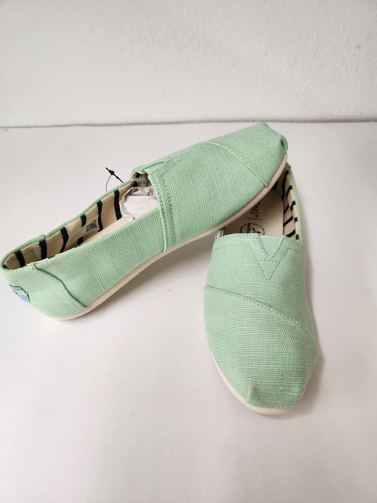 A pair of mint green canvas slip-on shoes, likely from Toms, displayed on a white surface against a white wall.