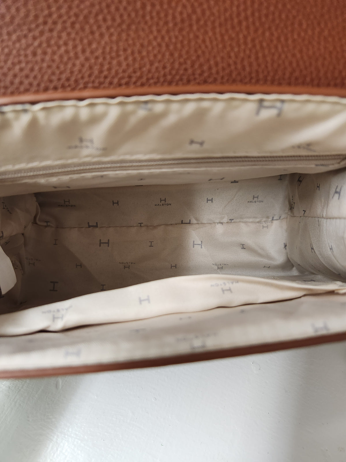 Close-up of the interior of a brown textured leather bag, showing a cream-colored lining with a repeating small H logo pattern.