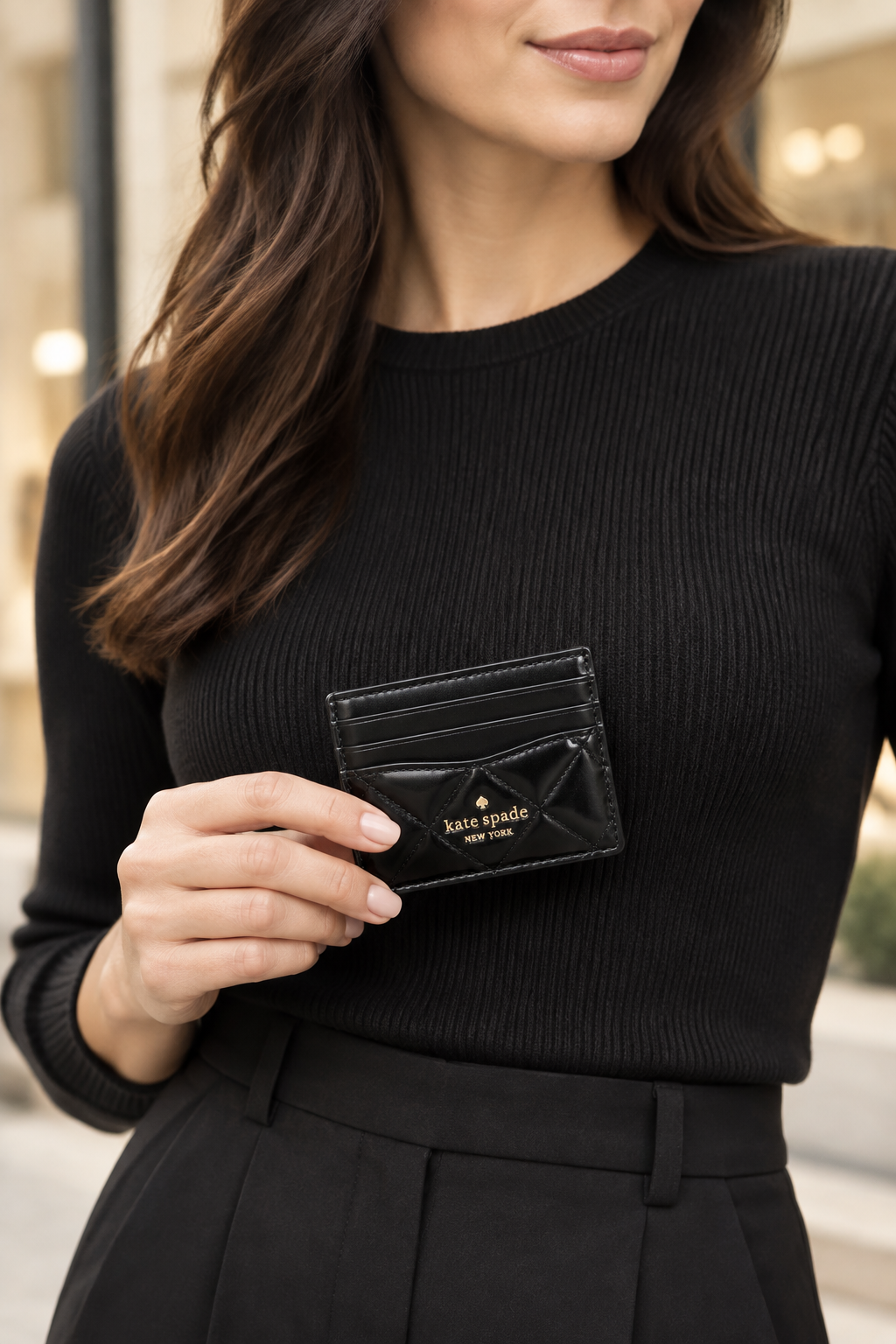 A woman in a black ribbed long-sleeve top holds a black leather card holder wallet by Kate Spade New York. The wallet features a prominent bow detail and gold logo lettering, set against a softly blurred outdoor background.