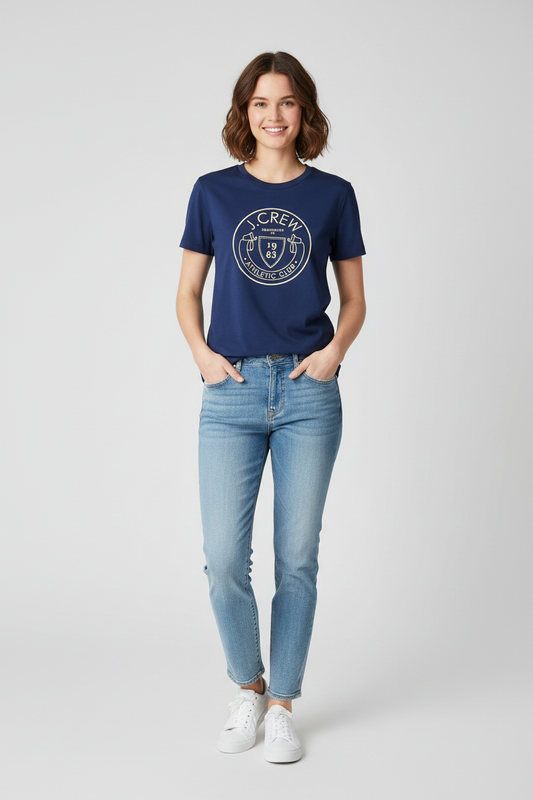 A woman modeling a navy blue crewneck t-shirt with a white circular graphic on the chest, paired with light-wash blue jeans and white sneakers, against a light gray studio background.