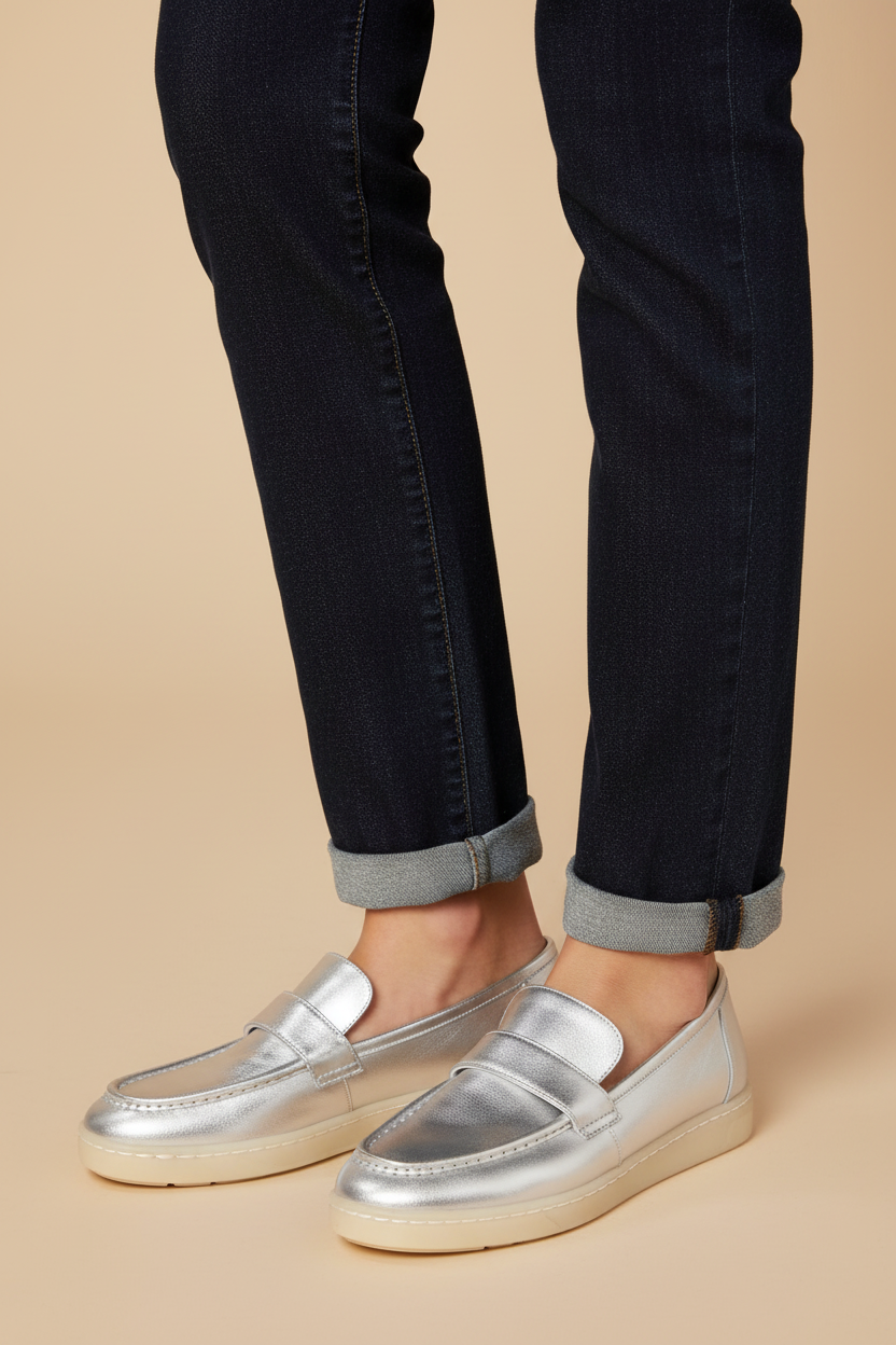 A person wearing shiny metallic silver loafers with dark cuffed jeans, shown from the knees down against a plain beige studio background.