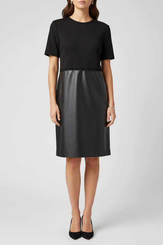 A woman modeling a black dress with a short-sleeve knit top and a smooth faux-leather skirt, paired with black heels, against a light gray studio background.