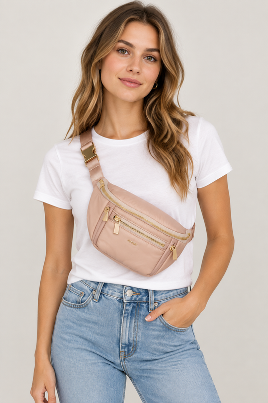 A woman wearing a light pink faux-leather crossbody fanny pack with gold-tone hardware, styled with a white t-shirt and light blue jeans against a light gray studio background.