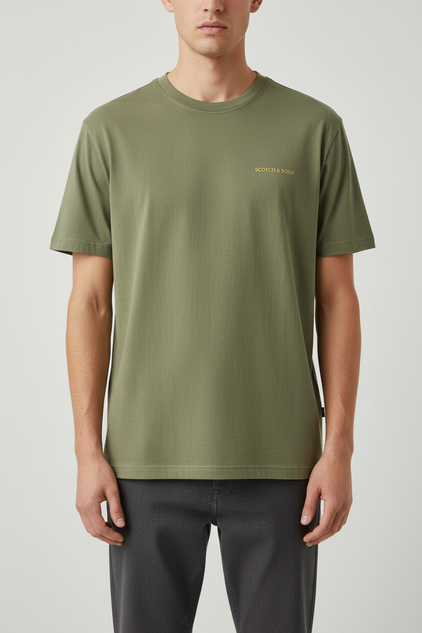 A man wearing an olive green short-sleeve crewneck t-shirt with a small gold 'BOTMONE BAGS' logo on the left chest, paired with dark gray pants, against a light gray studio background.