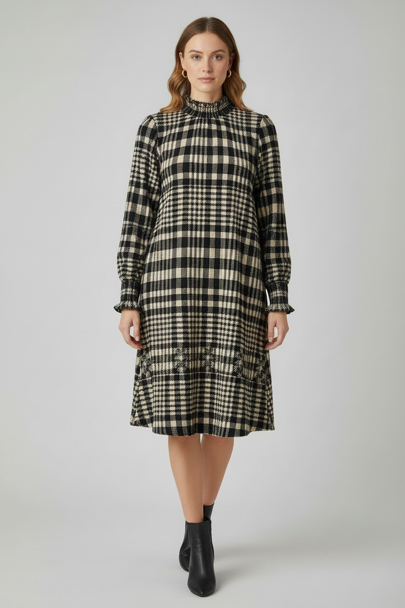 A woman modeling a long-sleeve black and beige plaid midi dress with a high gathered neckline and ruffled hem, paired with black ankle boots, against a light gray studio background.