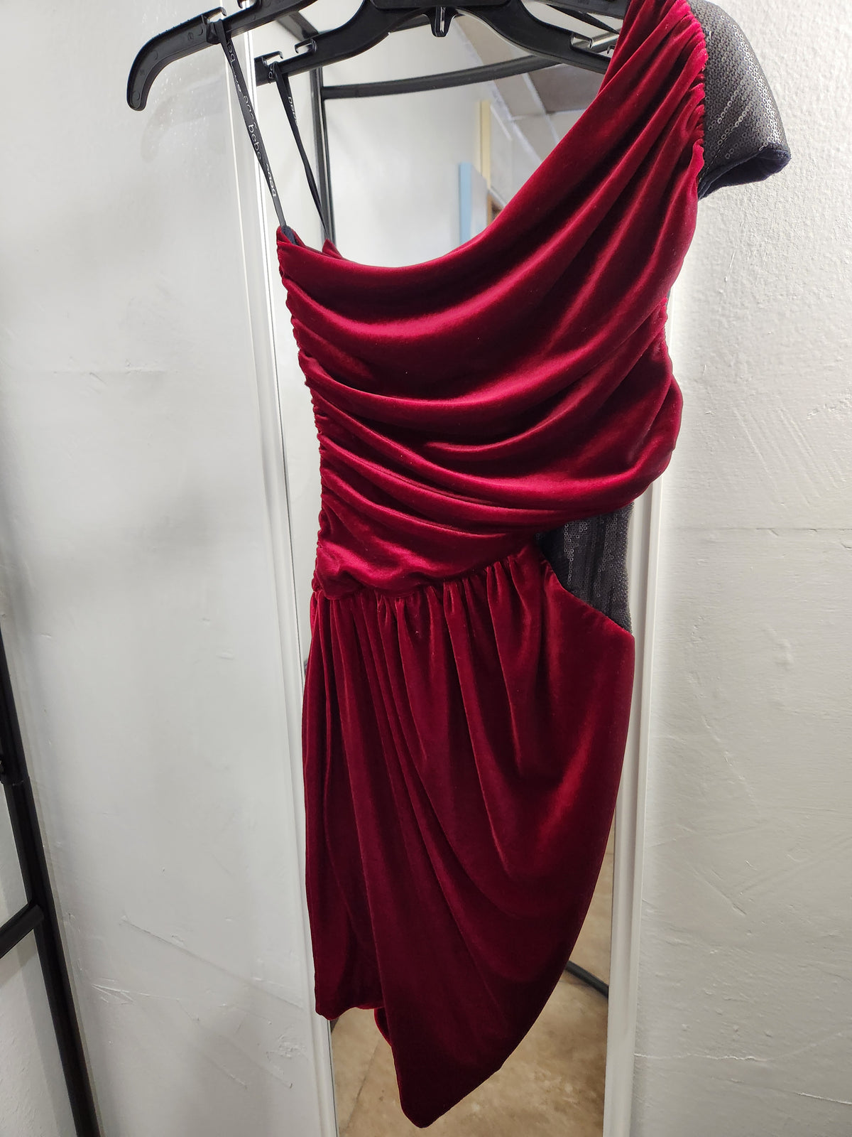 A deep red velvet one-shoulder dress hanging on a black hanger against a white wall.