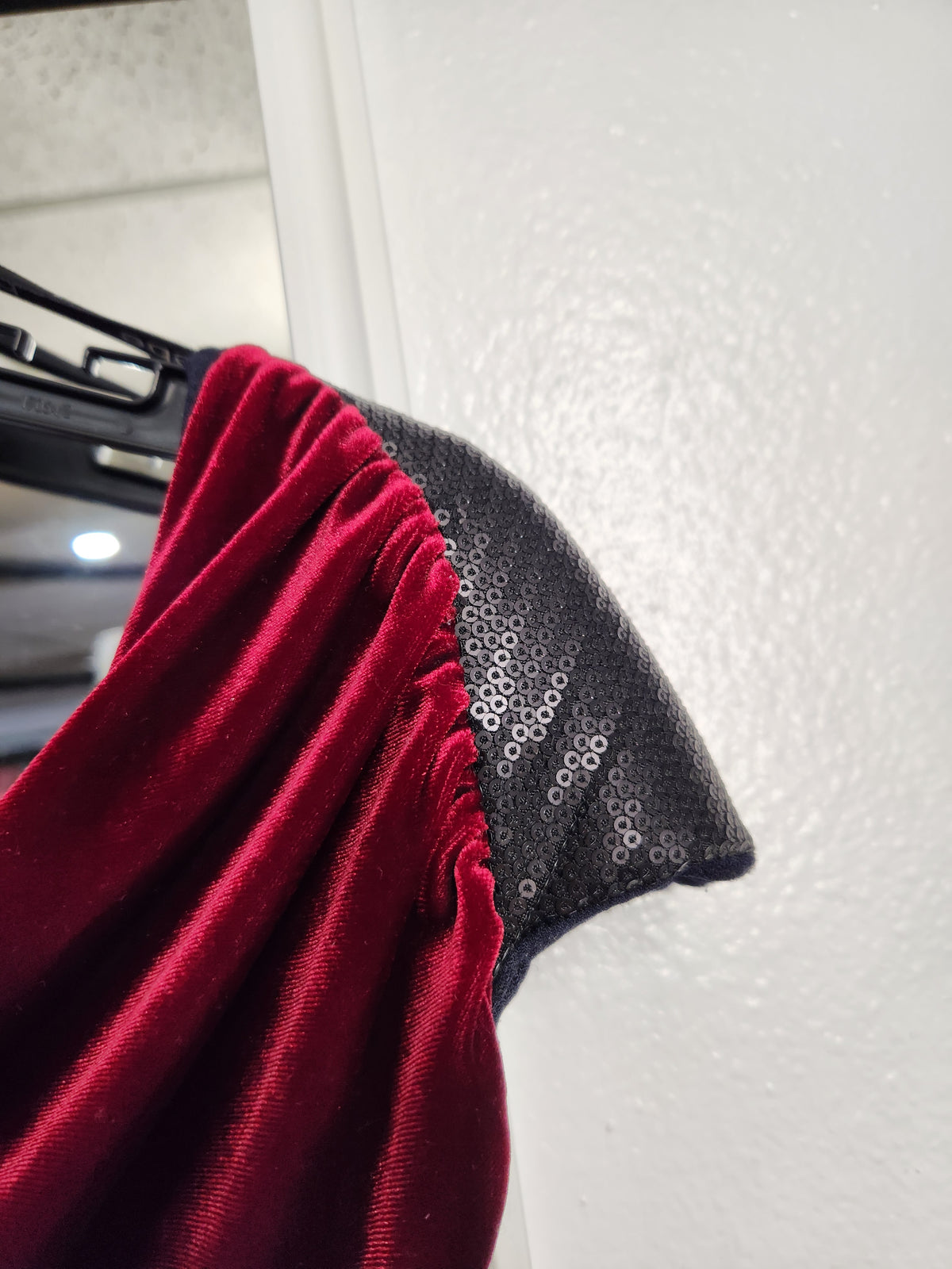 A close-up of a garment featuring a deep red velvet fabric and a black sequined fabric panel, hanging on a black hanger against a white textured wall.