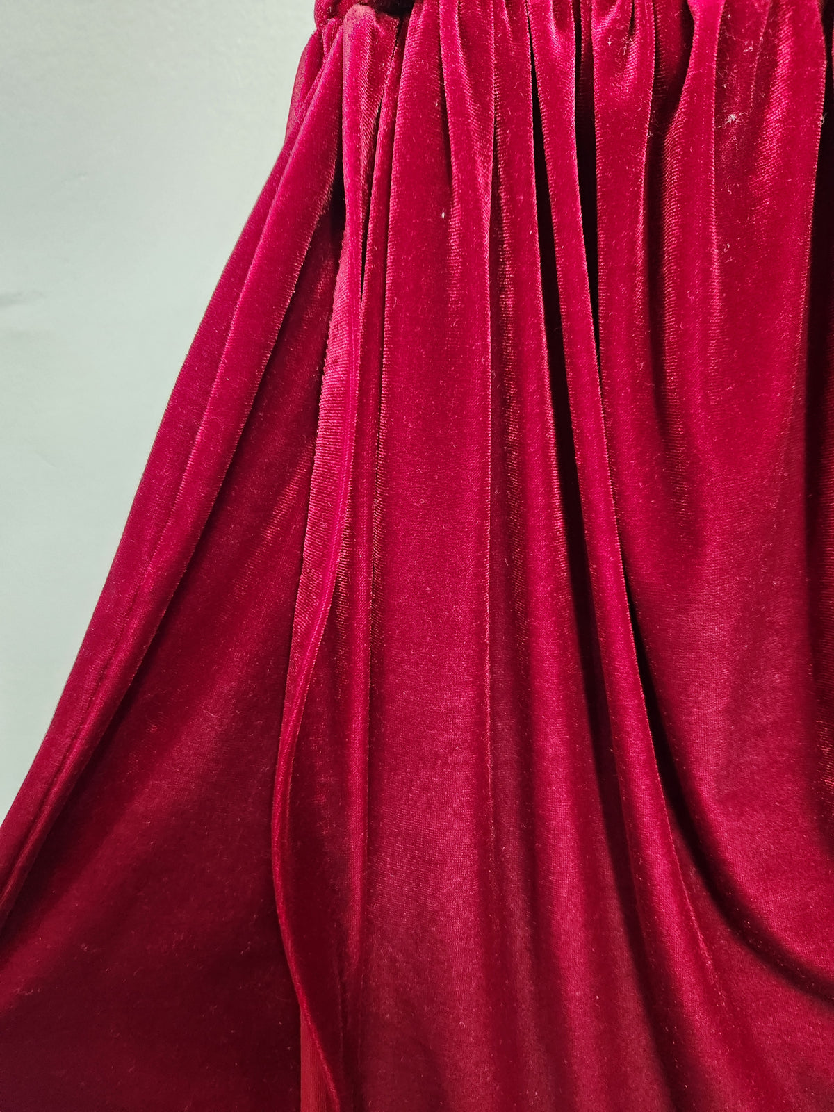 A close-up of draped deep red velvet fabric with soft folds and a smooth, plush texture against a plain light background.