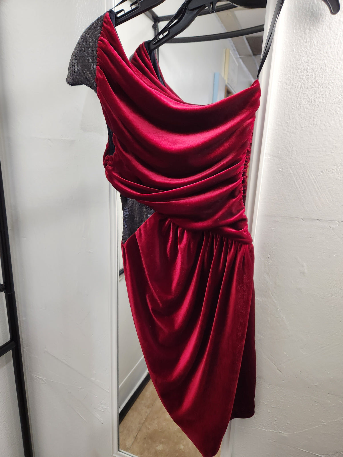 A deep red velvet one-shoulder dress with ruched draping details, hanging on a black hanger against a white wall.