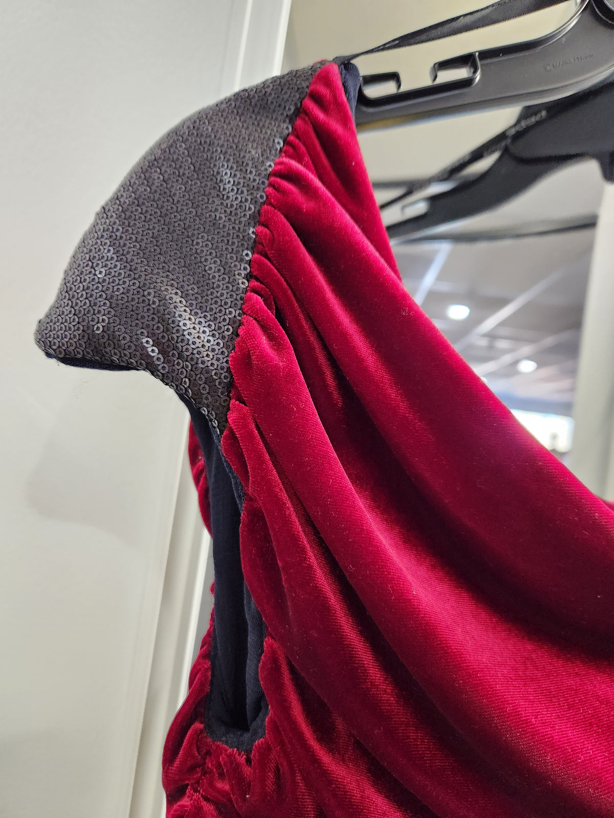A close-up of a red velvet garment draped over a black hanger, showing a dark gray sequined shoulder panel and gathered elastic details.