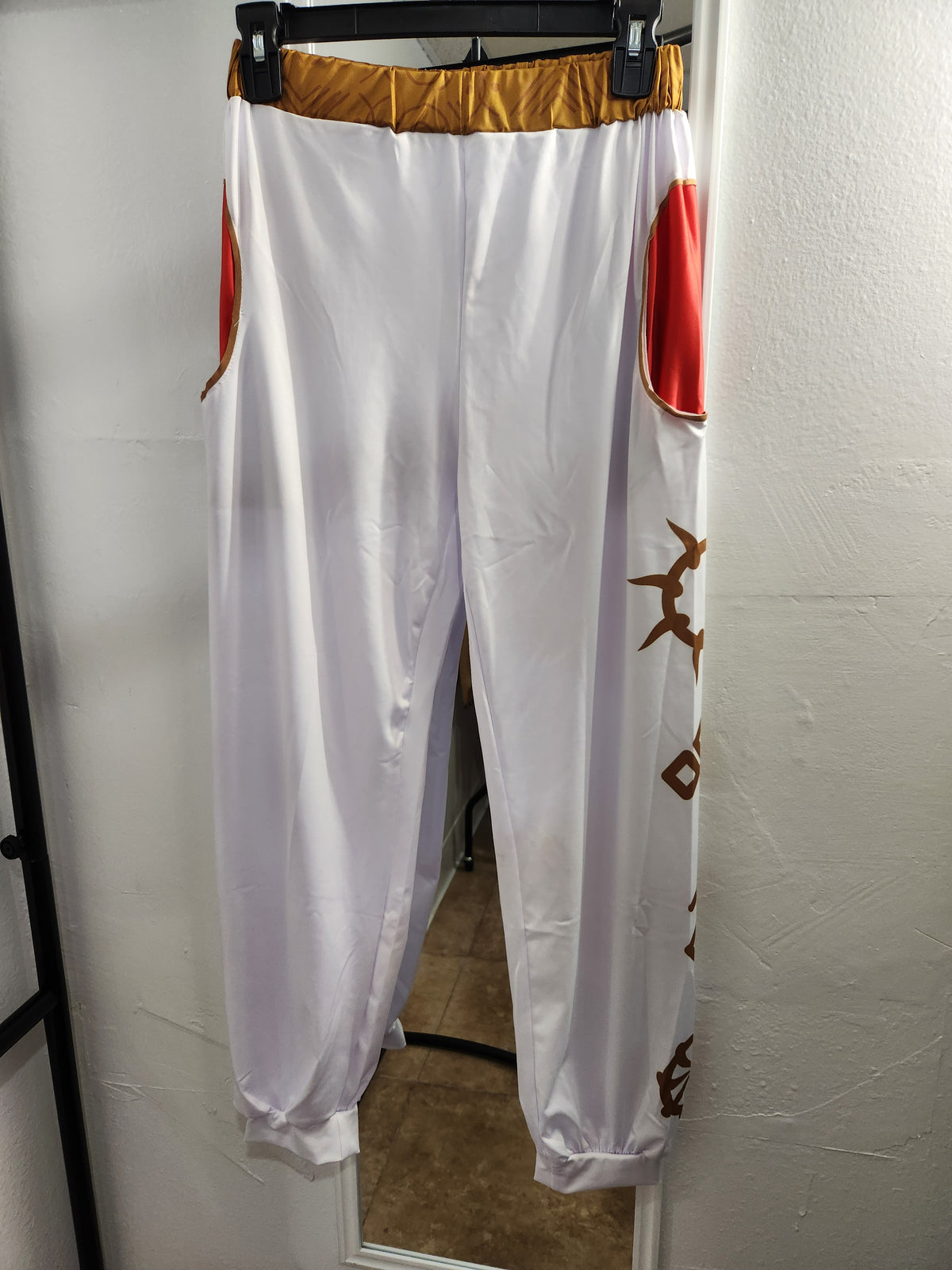 White costume pants with gold and red accents, hanging on a black hanger against a white wall.