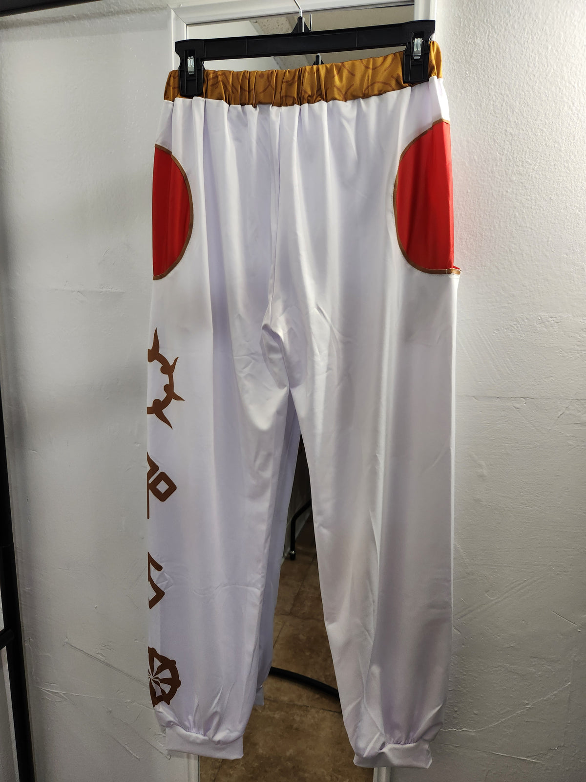 White pants with red circular patches on the upper legs, a gold elastic waistband, and brown geometric patterns down one leg, hanging on a black hanger against a white wall.