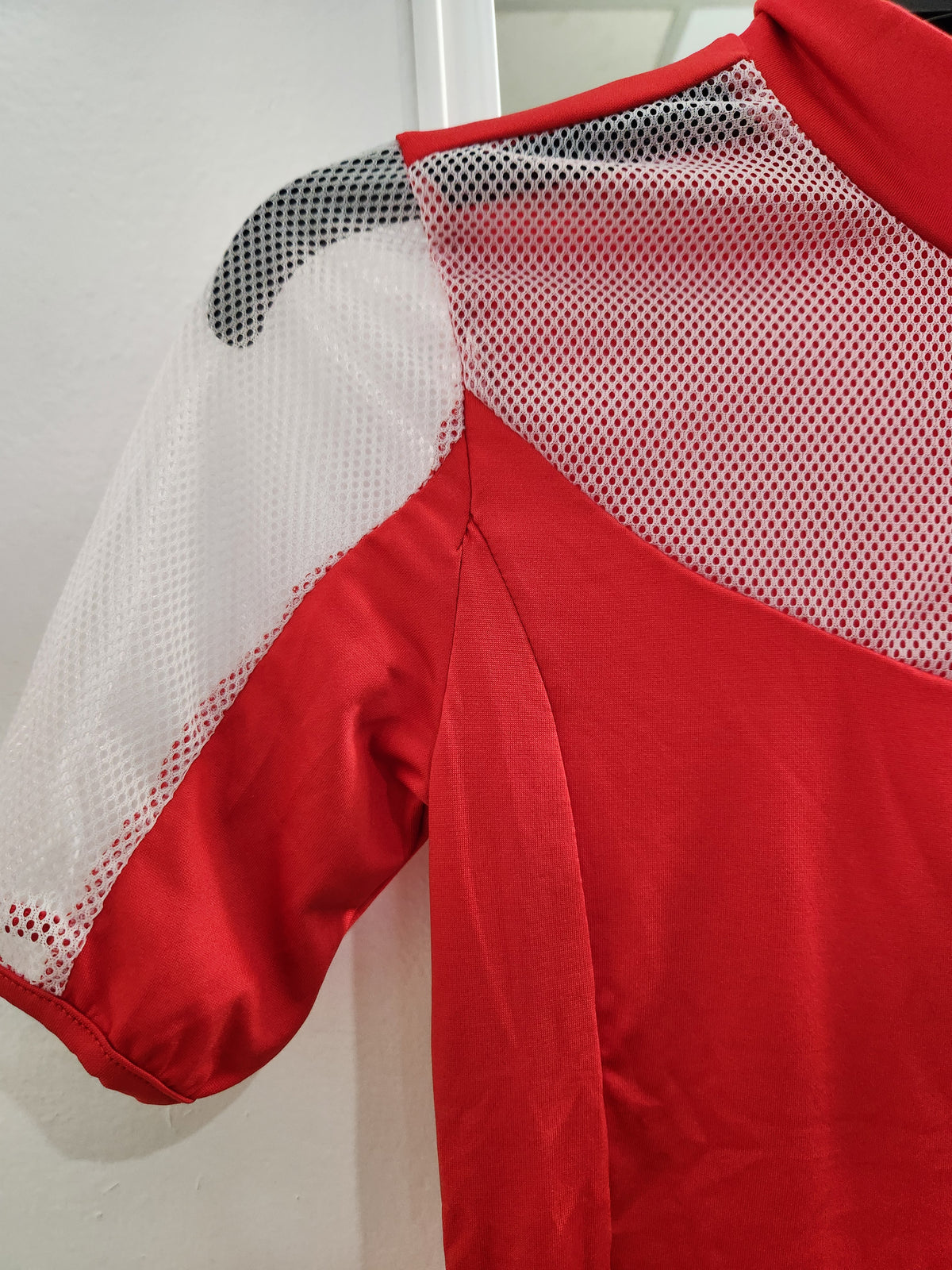A close-up of a red athletic jersey with white mesh shoulder panels and a black shoulder patch, shown against a plain light background.