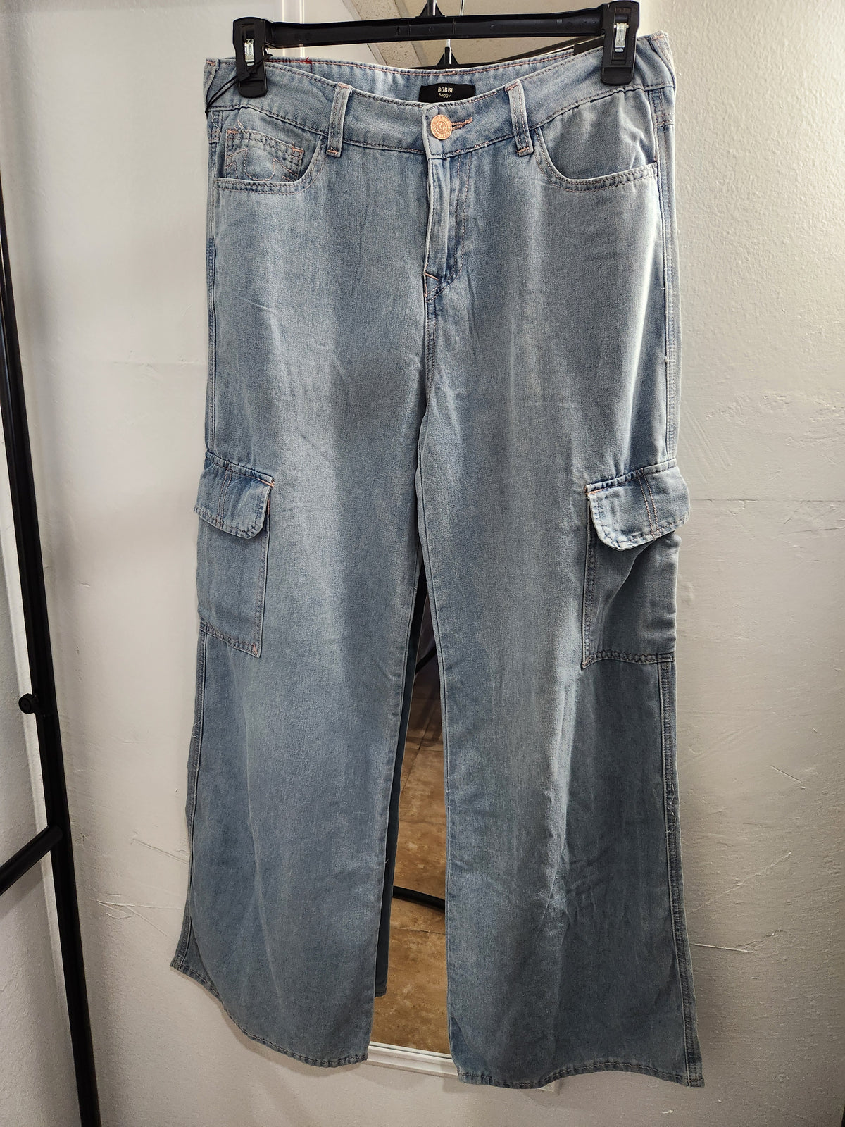 Light blue denim cargo pants with a wide-leg fit, hanging on a black hanger against a white wall by True Religion