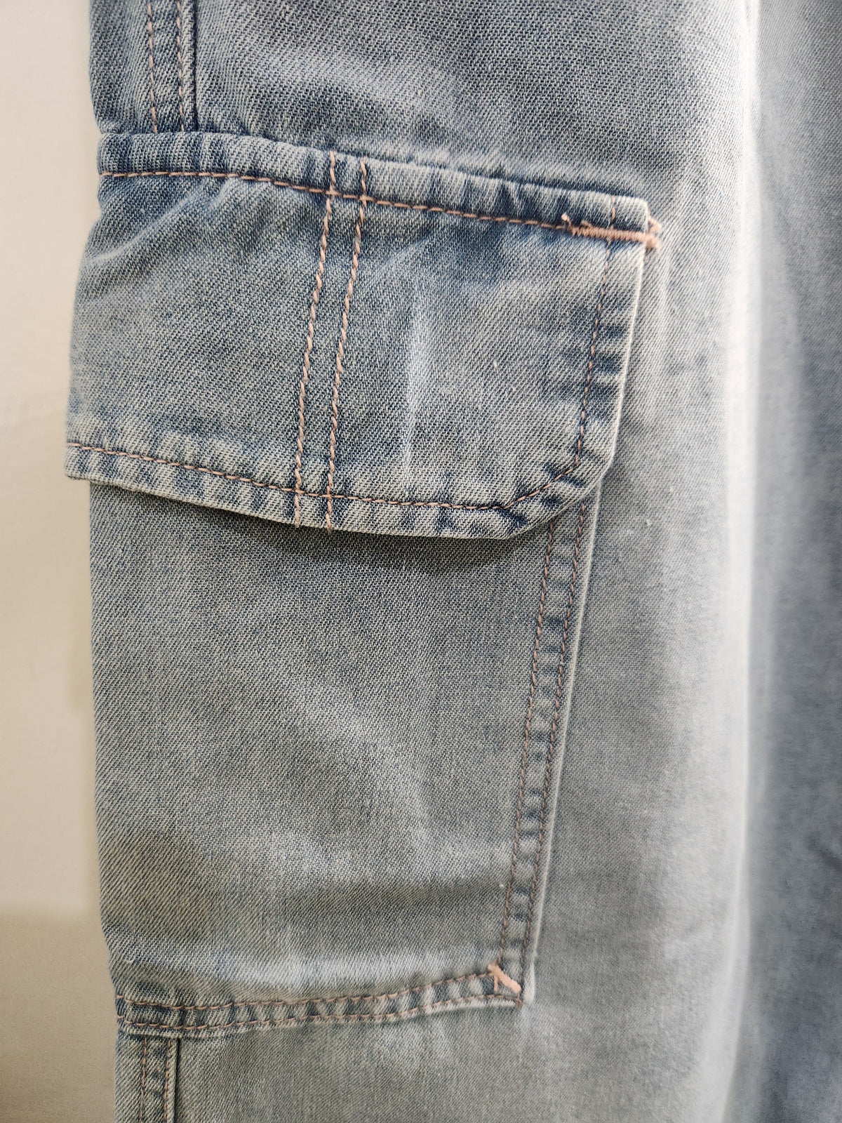 Close-up of a light blue denim garment, showing a flap pocket with contrast stitching.