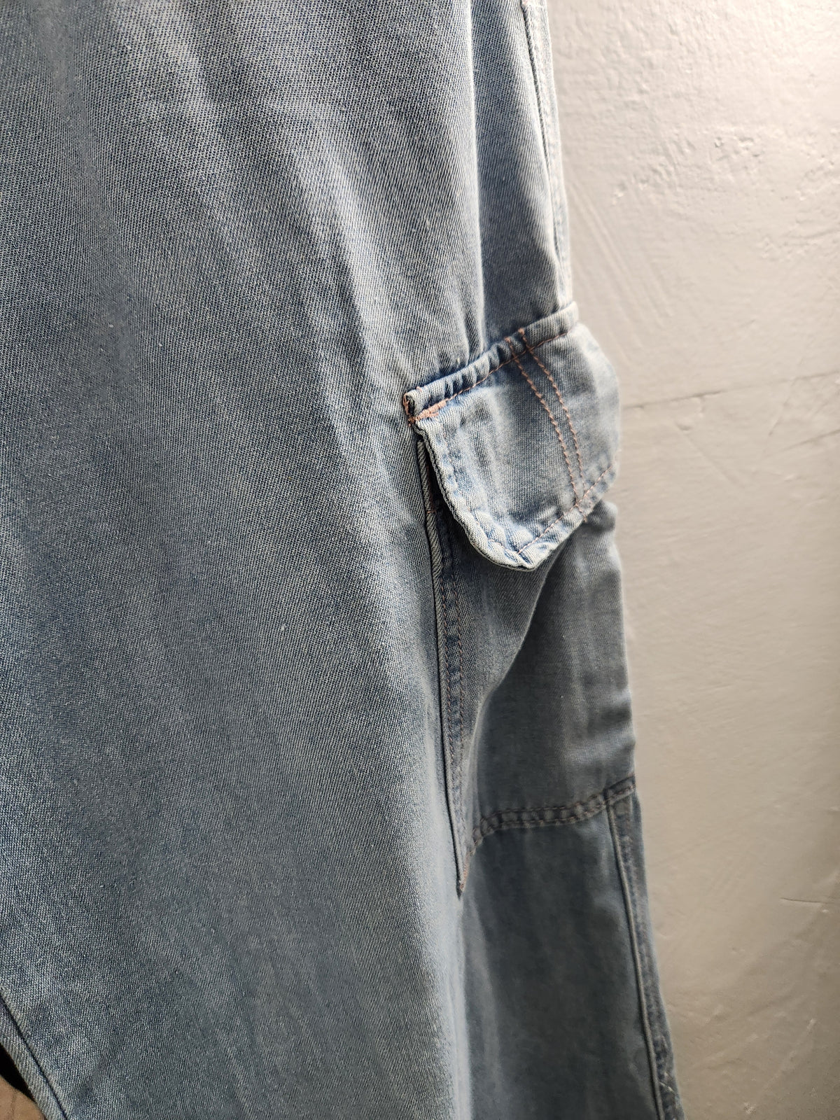 A close-up of a light blue denim garment, likely a dress or long skirt, featuring a side cargo pocket and a textured off-white wall in the background.