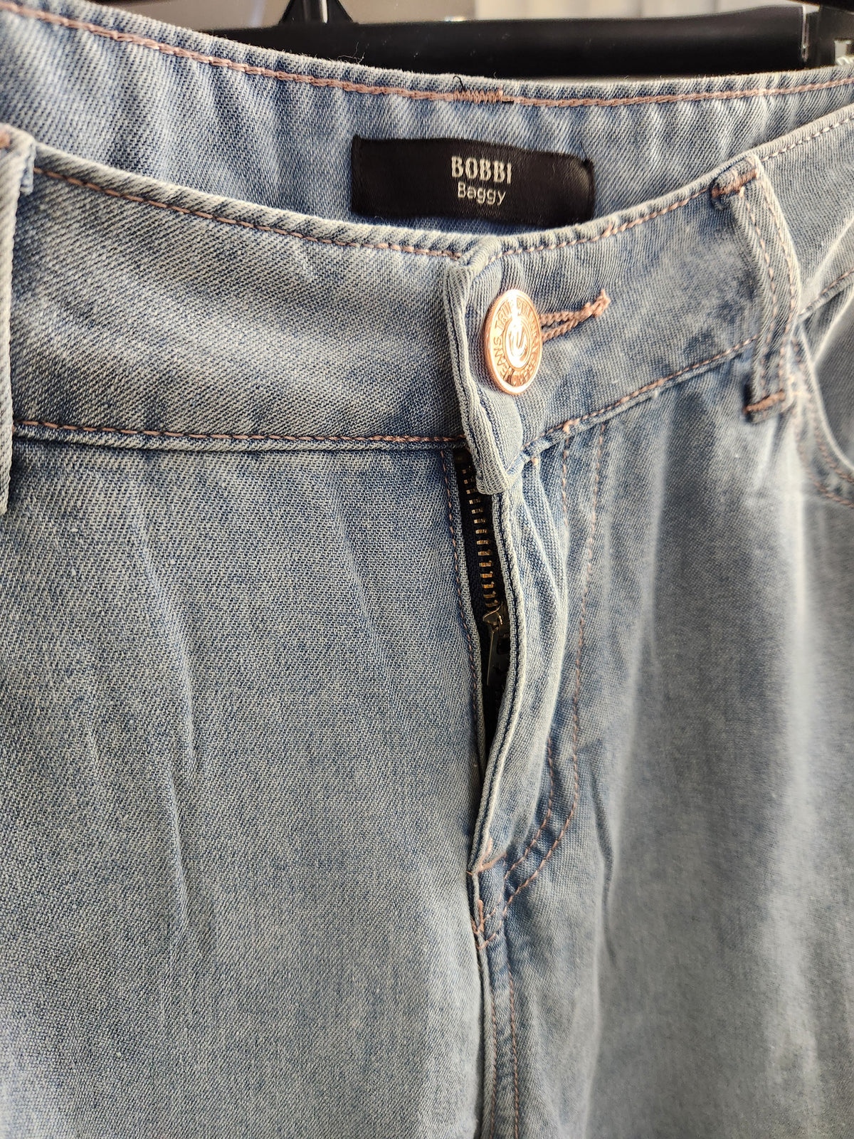 Close-up of a pair of light-wash blue denim jeans hanging on a black hanger, showing the waistband, button, zipper, and a black BOBBI Baggy brand label.