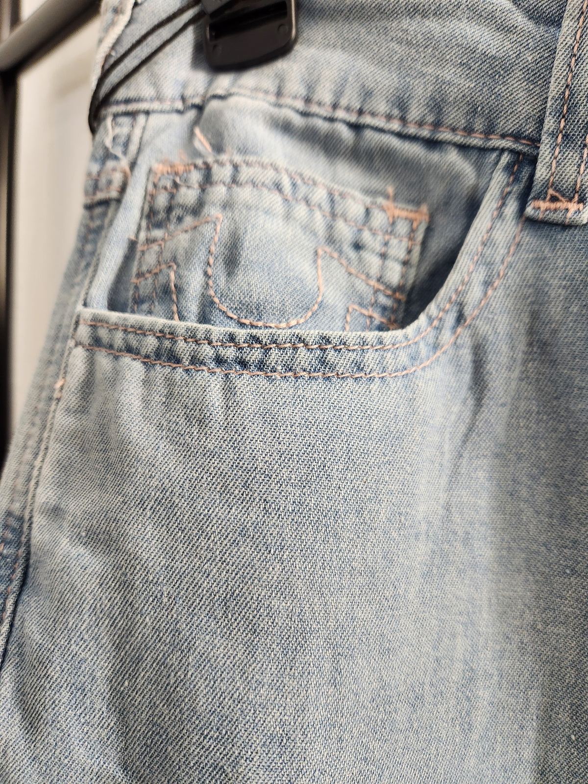 Close-up of a light blue denim pair of jeans showing the back pocket with contrast orange stitching and a black belt loop.