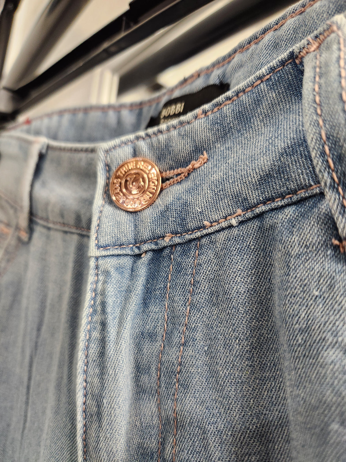 Close-up of a pair of blue denim jeans showing the waistband, button, and stitching details. A black label inside the waistband reads BOBBI.
