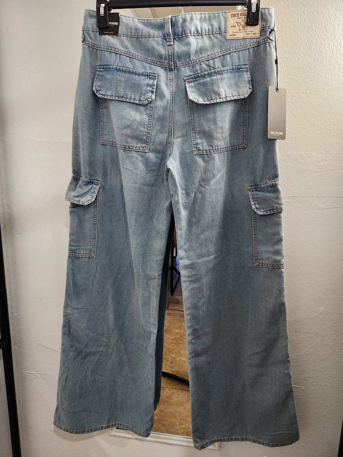 Light blue denim cargo pants with a faded wash, hanging on a black hanger against a white wall. The back view shows two large flap pockets on the upper back, side cargo pockets, and a hangtag attached.