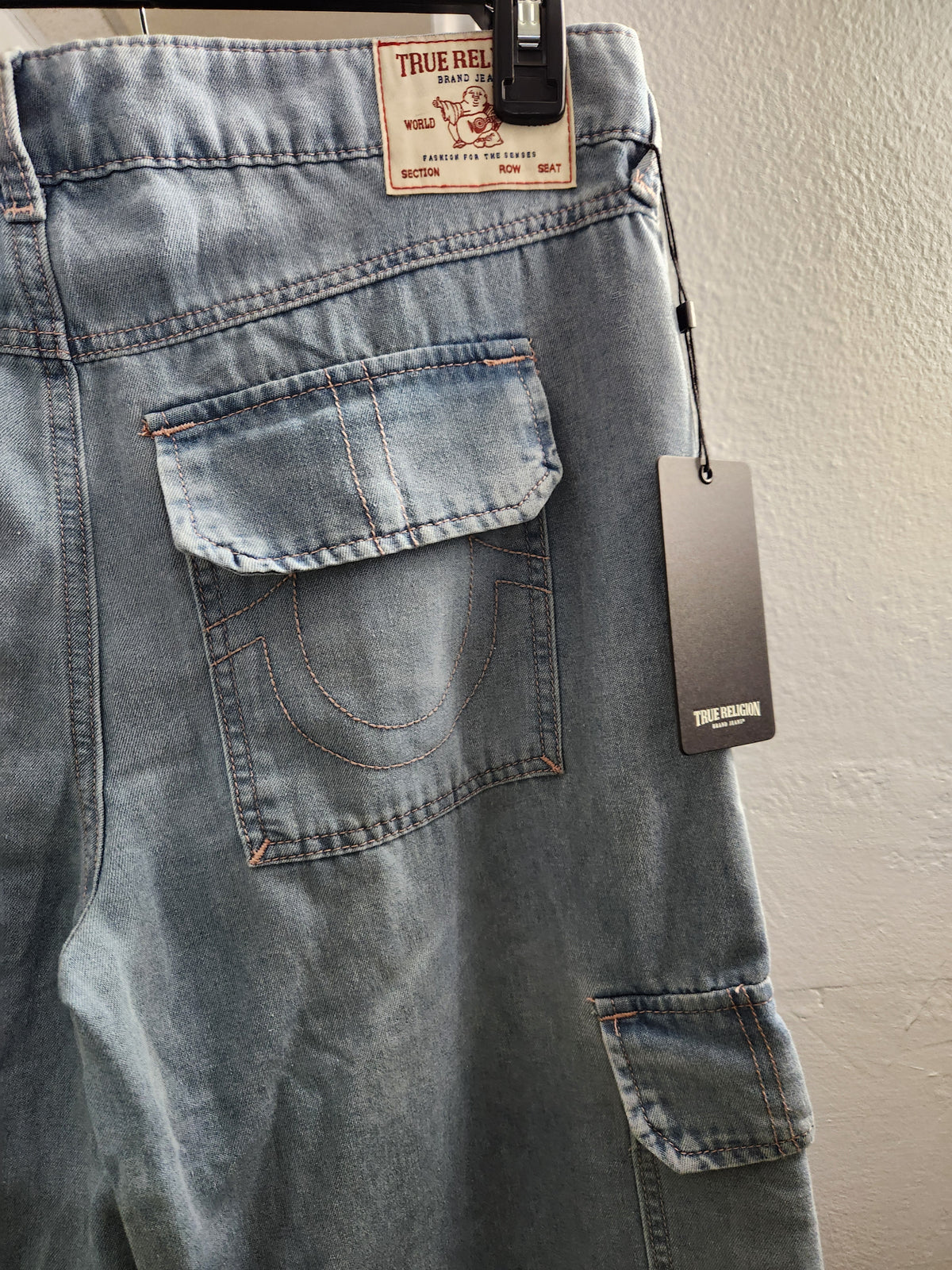Light blue denim cargo pants from True Religion, shown hanging on a black hanger against a textured off-white wall. The back pocket and a hangtag are visible.