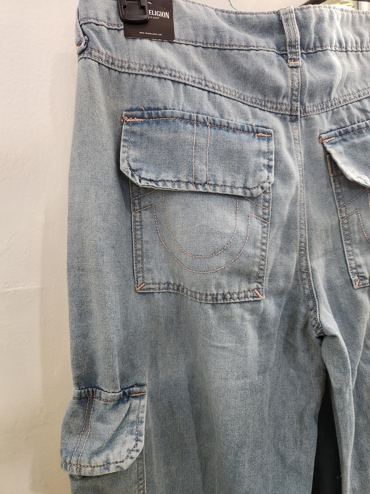 Close-up of the back waistband and cargo pockets of light-wash blue denim jeans, showing a dark brown brand label that reads True Religion