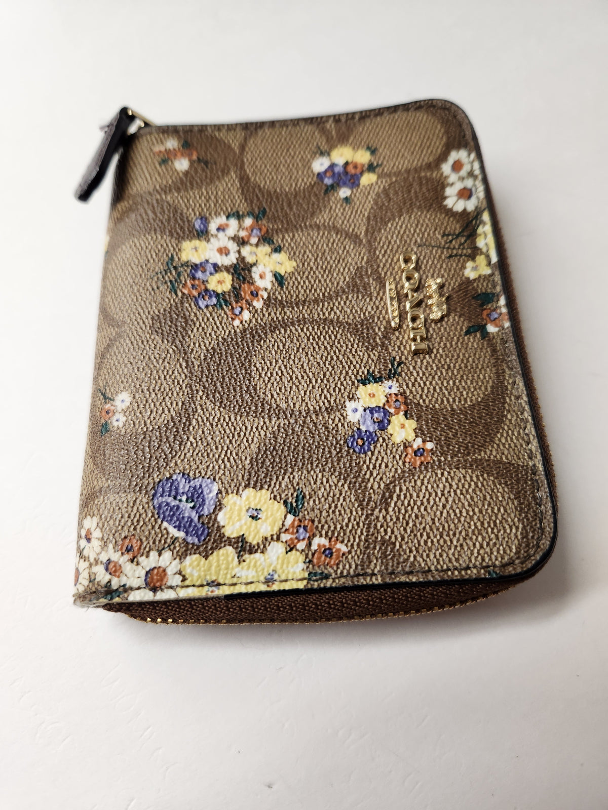 A small zip-around wallet in brown coated canvas with the signature Coach monogram and scattered floral prints, plus a gold-tone Coach logo on the front, photographed on a plain light background.
