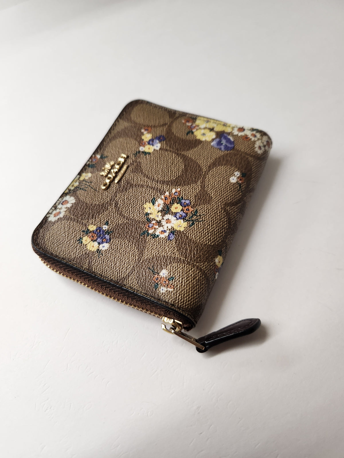 A small zip-around wallet in brown monogram canvas with a floral print, featuring a gold-tone Coach logo clasp and a dark brown zipper pull, photographed on a light off-white background.