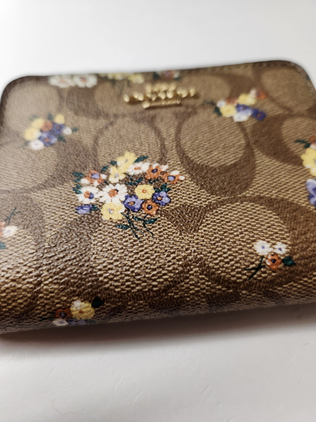 A close-up of a Coach wallet in the brand's signature brown monogram canvas, decorated with small colorful floral bouquets in yellow, white, and purple. A gold-tone Coach logo is visible near the top edge.