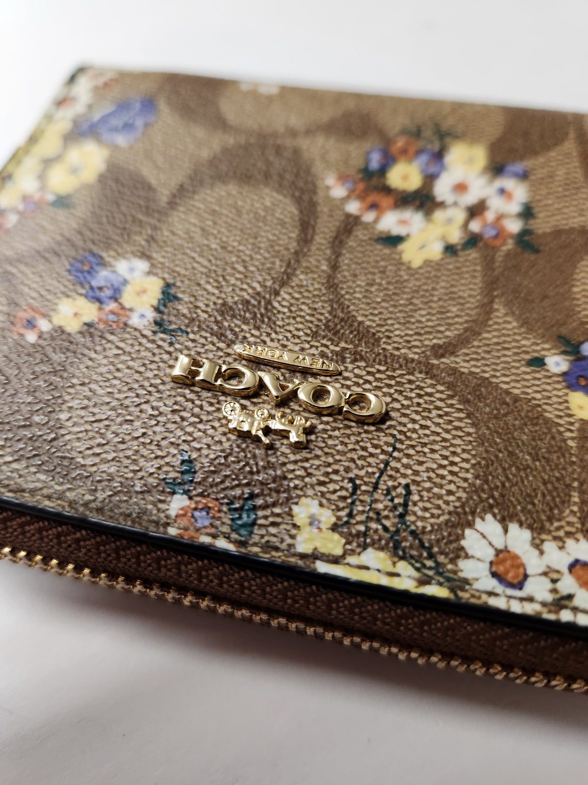 A close-up of a Coach wallet in their signature brown monogram canvas with a colorful floral print and gold-tone logo hardware.