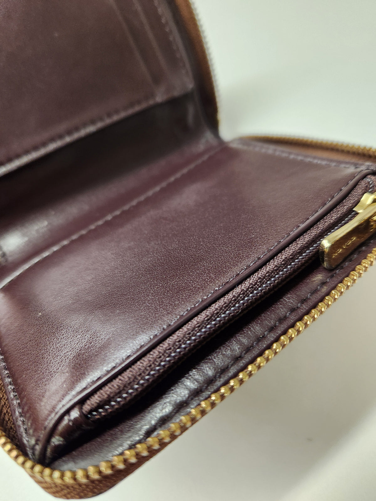 Close-up of an open brown leather wallet showing interior card slots, stitching, a zip compartment, and a gold-tone zipper pull on a light background.