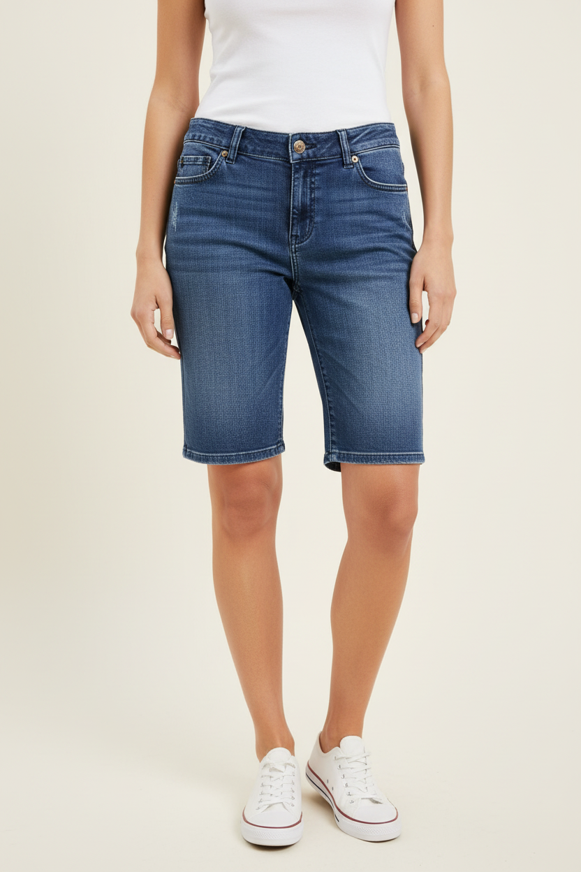 A model wearing a pair of medium-wash blue denim shorts with a white tank top and white sneakers, shown from the waist down against a light beige studio background.