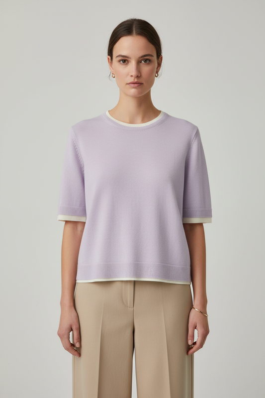 A woman modeling a light lavender short-sleeve knit top with cream ribbed trim at the crew neck, sleeve cuffs, and hem, paired with beige wide-leg trousers, against a neutral light gray background.