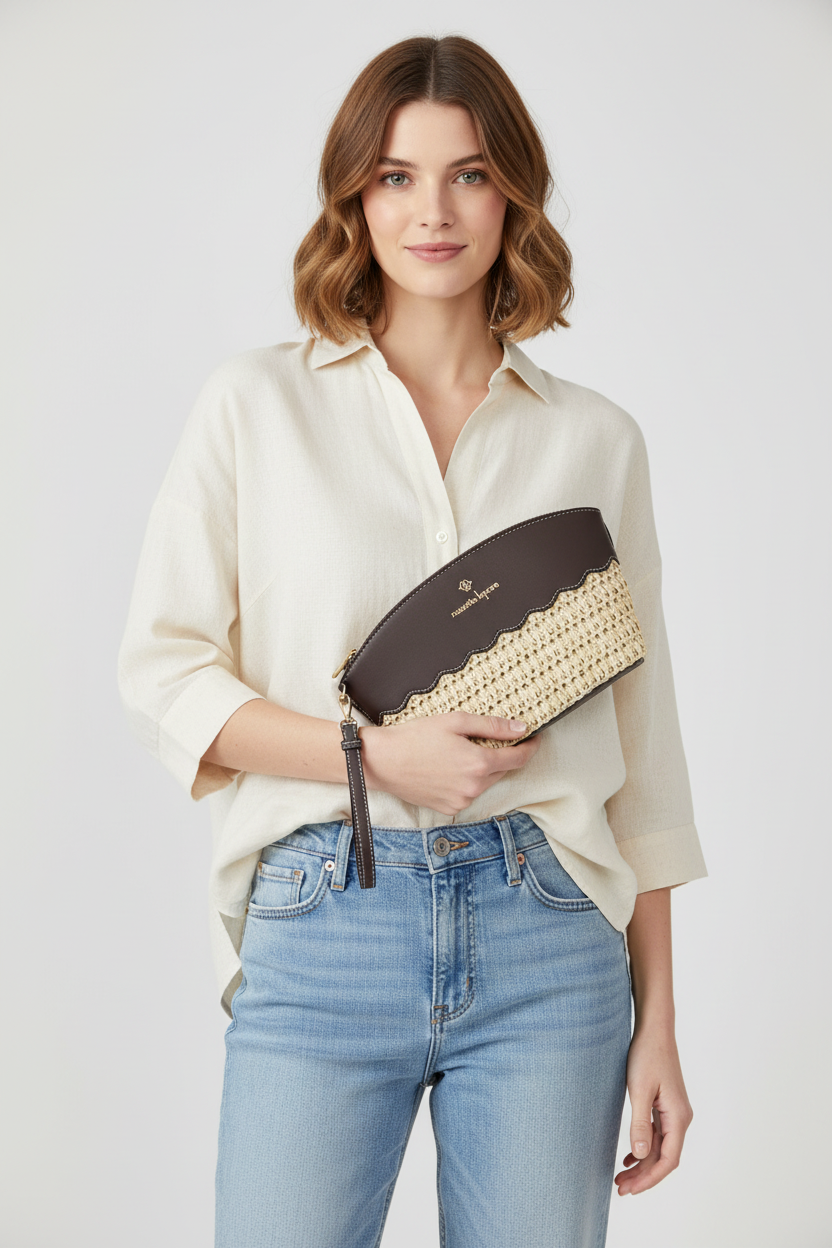 A woman wearing a cream button-down shirt and light blue jeans holds a dark brown and beige woven clutch bag with a gold logo.