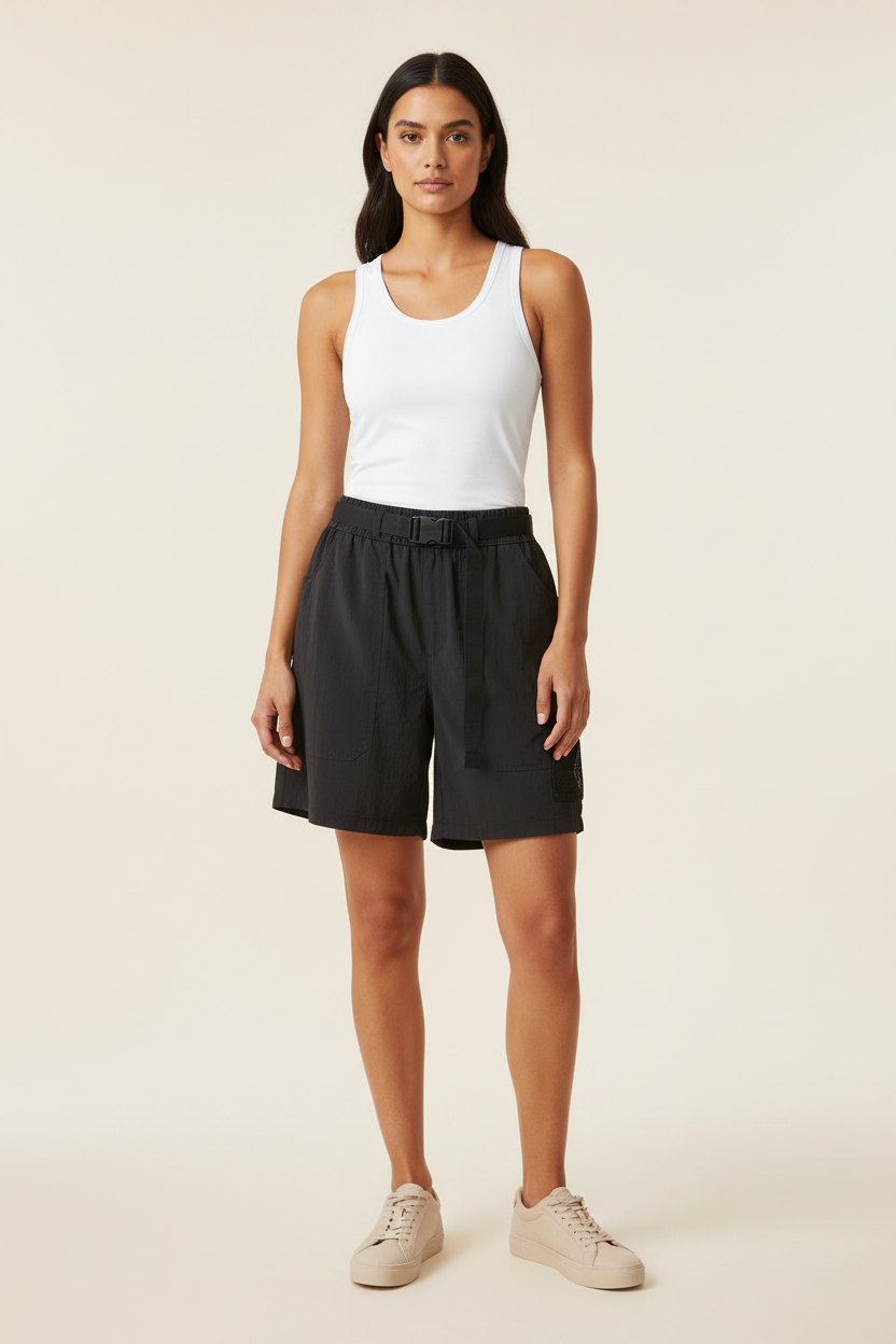 A woman modeling black athletic shorts with a matching black belt, paired with a white tank top and beige sneakers, against a solid light beige background.