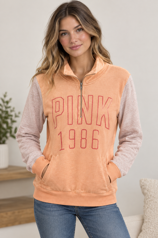 A woman wearing a PINK 1986 quarter-zip sweatshirt with a peach-colored body and heathered light pink sleeves, paired with blue jeans in a brightly lit indoor setting.