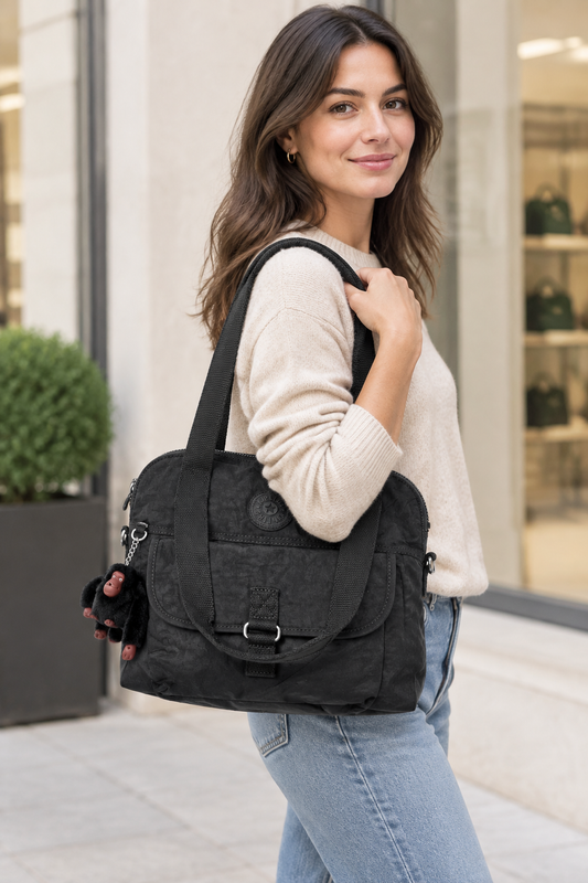 A woman wearing a cream knit sweater and light blue jeans carries a large black textured tote bag with a front flap and buckle closure, standing outdoors in front of a storefront.