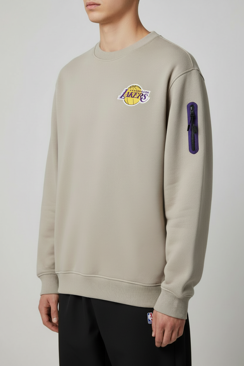 A man wearing a beige Los Angeles Lakers crewneck sweatshirt with a purple and yellow logo on the chest and a vertical purple pocket detail on the sleeve, paired with black pants.