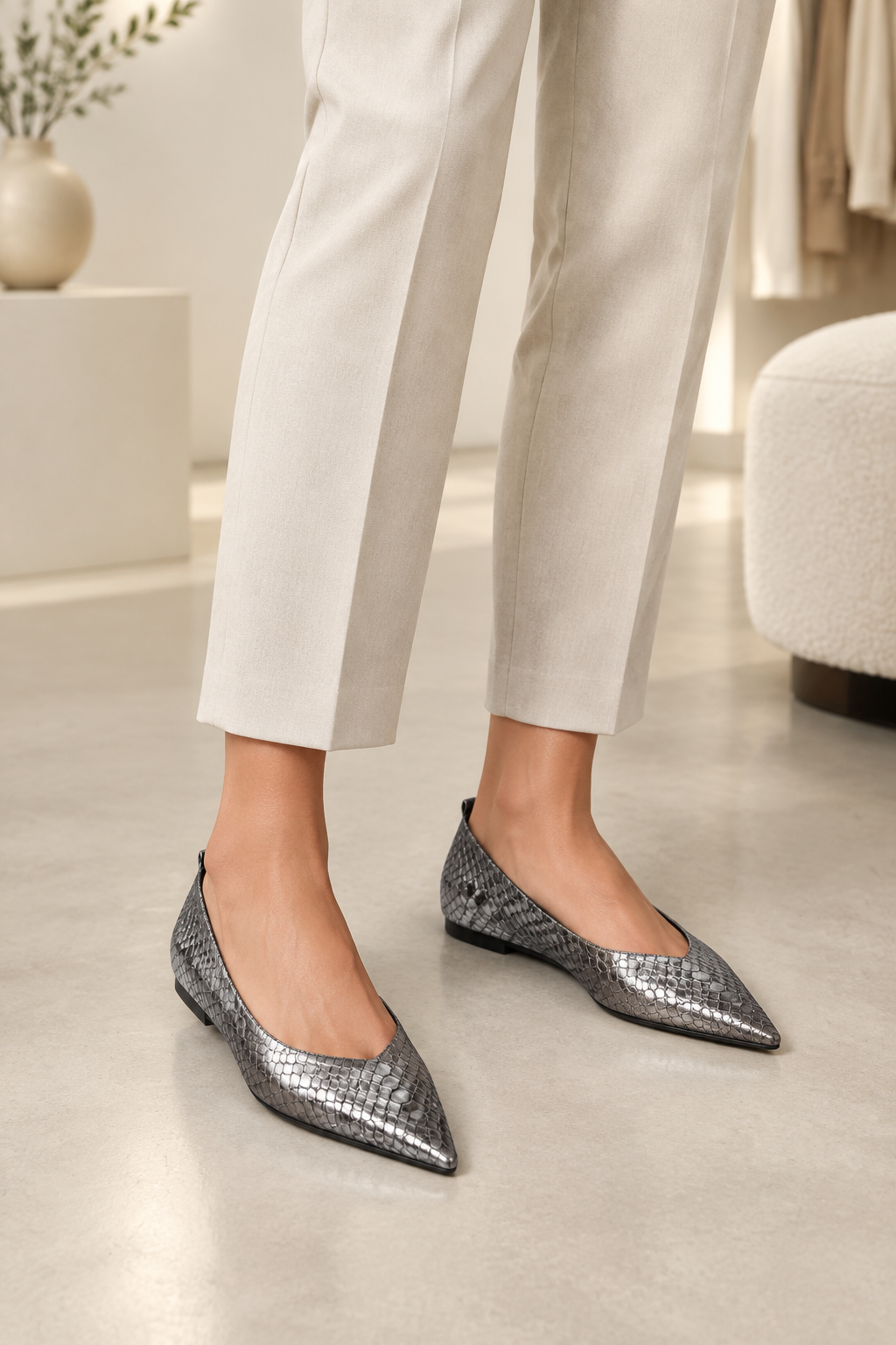 A person wearing pointed-toe metallic silver flats with a snakeskin texture, paired with light beige cropped trousers, standing in a bright, modern indoor setting.
